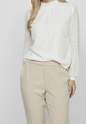 Kobieta nosząca białą, teksturowaną bluzkę z długim rękawem i marszczonymi mankietami, połączoną z beżowymi, dopasowanymi spodniami z wysokim stanem.