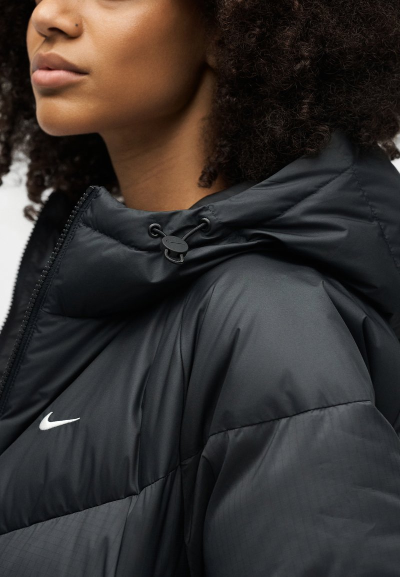 Nahaufnahme einer Person mit lockigem Haar, die eine schwarze Nike-Steppjacke mit Kapuze und verstellbarem Kordelzug trägt.