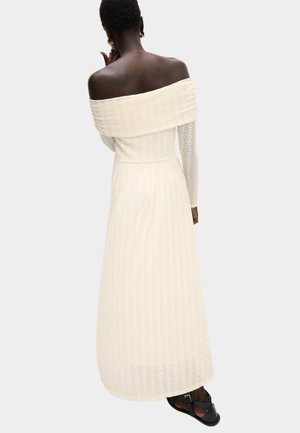 Kobieta widziana od tyłu, ubrana w kremową, długą sukienkę bez ramion o fakturowanej tkaninie i czarne sandały, stojąca na jasnym, gładkim tle.