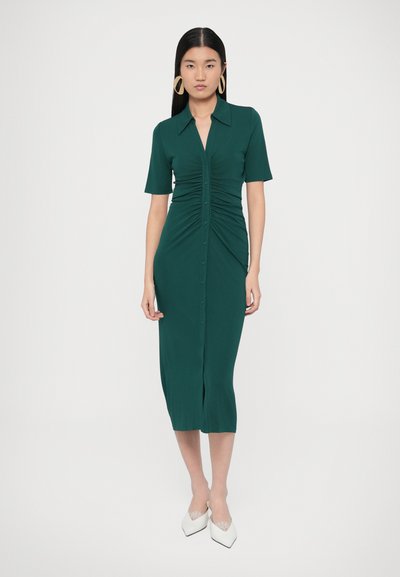 Tyrkysov�é košeľové šaty s krátkymi rukávmi, nariasenými textúrami na prednej strane a dĺžkou po polovicu lýtok. Zladené s bielymi špicatými topánkami.