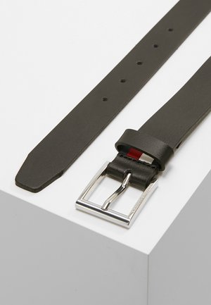 Zwarte leren riem met een zilverkleurige rechthoekige gesp, meerdere verstelgaten en een klein rood detail vlak bij de gesp, plat gelegd op een witte ondergrond.