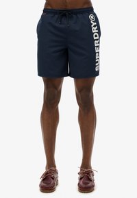 Shorts de bain bleu marine avec le logo "SUPERDRY" en blanc sur le côté. Ils possèdent une taille élastique avec un cordon de serrage et un matériau léger et lisse.