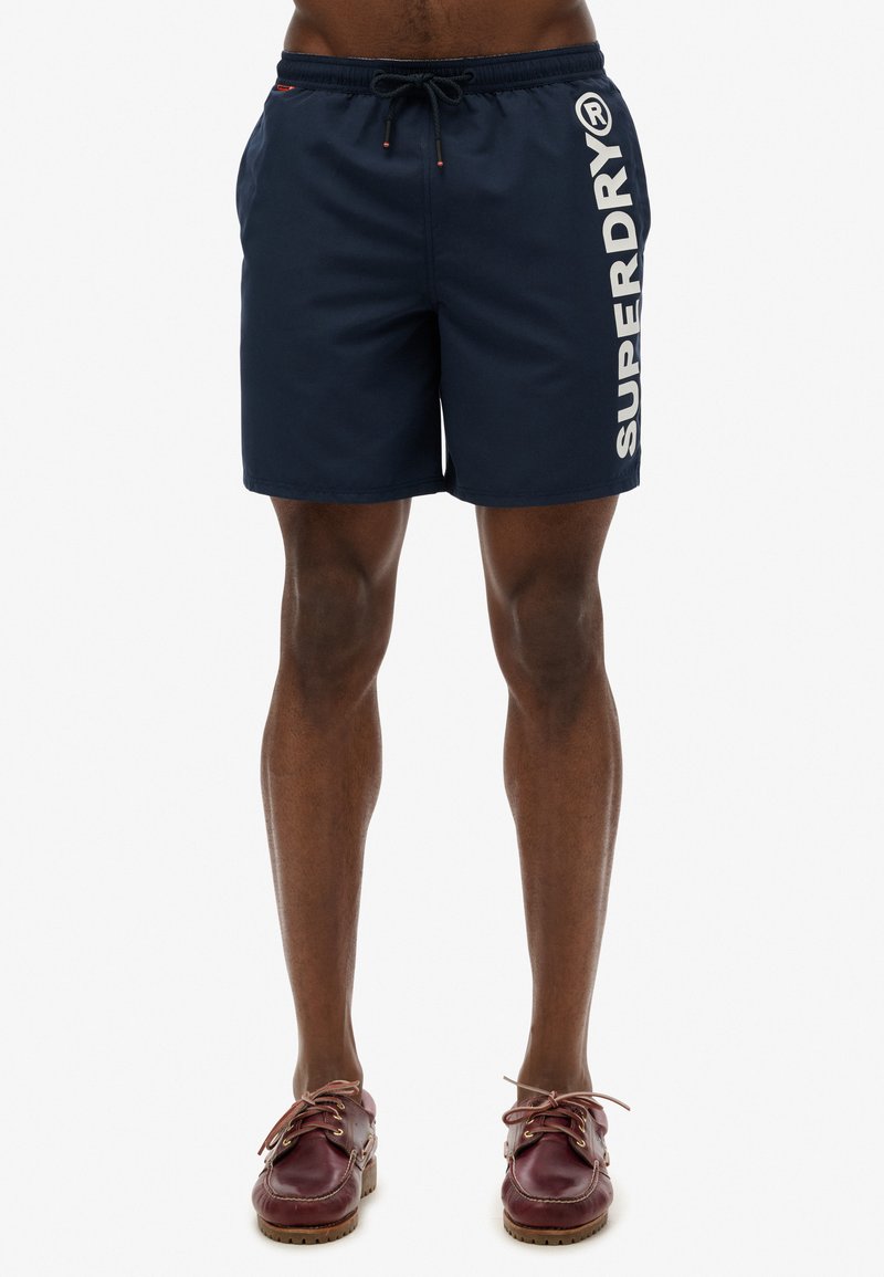 Shorts de bain bleu marine avec le logo "SUPERDRY" en blanc sur le côté. Ils possèdent une taille élastique avec un cordon de serrage et un matériau léger et lisse.