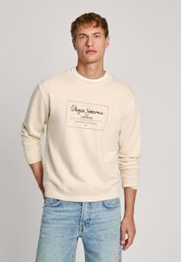 Felpa di colore beige chiaro con scollo rotondo, polsini e vita a costine. Presenta una stampa grafica scura con "Pepe Jeans London" sul davanti.