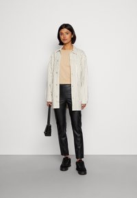 Camisa riscada em branco e bege sobre um top bege, calças de couro pretas e ténis pretos. A segurar uma carteira preta. Design minimalista.