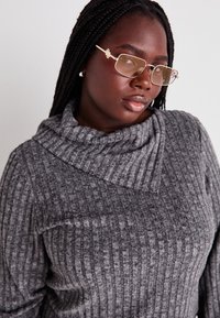 Pull gris à côtes avec un col cheminée, fabriqué dans un matériau texturé. L'individu a les cheveux tressés et porte des lunettes rectangulaires marron clair.