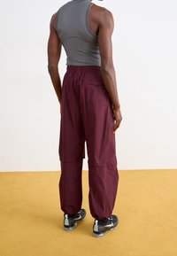 Bordeauxkleurige, losse broek met een elastische tailleband en zijzak, gecombineerd met een grijze tanktop en zwarte sportschoenen.