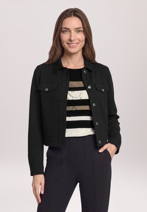 Femme aux cheveux bruns portant une veste noire boutonnée sur un haut rayé et un pantalon foncé, souriante, une main dans la poche.