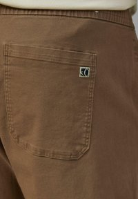 Bruine katoenen broek met een strak gesneden achterzak met een klein blauw-wit logo, gemaakt van een gladde, gestructureerde stof.