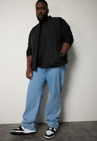 Schwarze, leichte Jacke über einem schwarzen Shirt, kombiniert mit hellblauen Jeans und schwarz-weißen Sneakers. Glattes Material mit lässigem Schnitt.