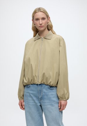 Frau trägt eine beige Reißverschlussjacke mit elastischem Saum und Bündchen, kombiniert mit hellblauen Jeans, stehend vor weißem Hintergrund.