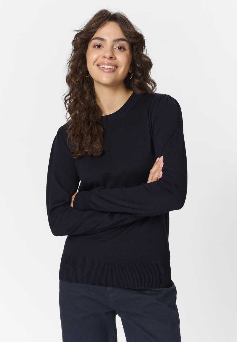 Tumši zils adīts džemperis ar garām piedurknēm, apaļu kakla izgriezumu un ribotu apakšmalu. Iespējams, ka tam ir smalka tekstūra visā virsmā un savākta pleca detaļa.