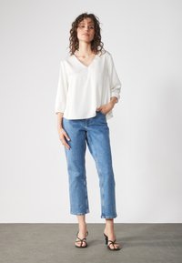 Hvit V-hals bluse med løse 3/4 ermer, paret med blå cropped jeans. Modellen har på seg svarte strappy sandaler. Ensfarget, glatt stoff.