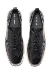 Un paio di scarpe lace-up in pelle nera con punte a forma di ala, dotate di suole in gomma bianche e beige, viste dall'alto, con il marchio "Cole Haan" all'interno.