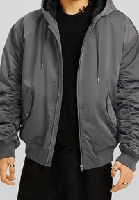 Grå bomberjacka med huva, elastiska ärmar och dragkedjestängning. Har rynkade ärmar och sidfickor; tillverkad av ett slitstarkt, glänsande material.