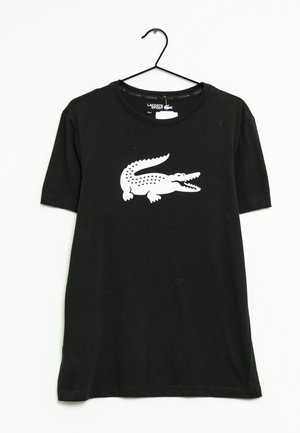 T-shirt nera a maniche corte di Lacoste Sport con un grande logo del coccodrillo bianco al centro del petto, appesa a una gruccia nera su uno sfondo bianco.