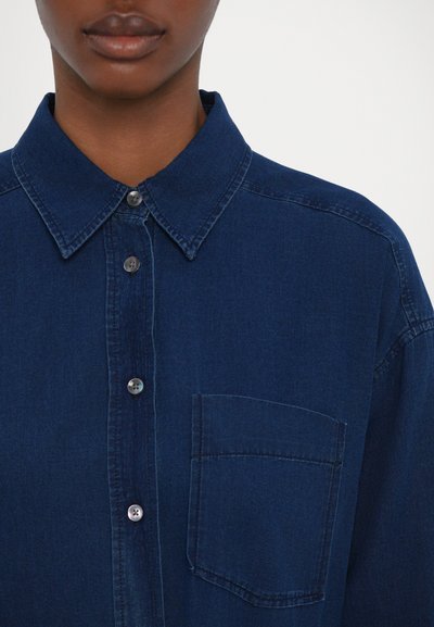 Mørkeblå denim skjorte med knapper, der har klassisk krave, sølvknapper og en brystlomme, samt en blød og glat tekstur.