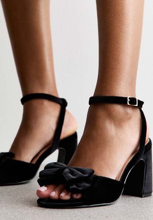 Svarta sammetsklackade sandaler med en rosettdetalj framtill, öppen tå-design och ankelrem med en metallspänne. Klack i blockstil.