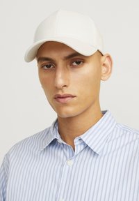 Hvid baseballkasket med en glat stoffinish, buet skygge og broderede detaljer. Kombineret med en lyseblå og hvid stribet skjorte.