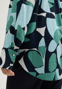 Chemise à manches longues en tissu texturé avec des motifs de feuilles abstraites en vert, bleu et marine. Elle dispose de poignets boutonnés et d'un ourlet incurvé.