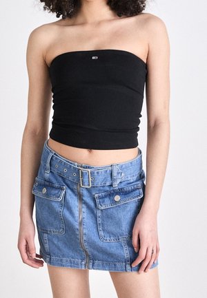 Femme portant un crop top noir sans bretelles et une mini-jupe en denim bleu avec fermeture éclair devant et ceinture, debout devant un fond uni.
