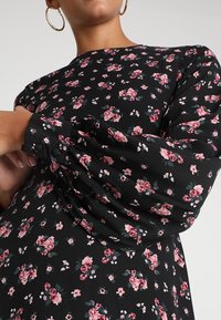 Květinové šaty s černým pozadím, zdobené růžovými a zelenými květinov ými vzory, s dlouhými nabíranými rukávy a knoflíkovými manžetami z jemné tkaniny.