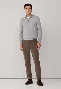 Grå tröja med dragkedja i halsen och långa ärmar, kombinerad med bruna byxor. Modellen har mörka skor. Enkel design, slät textur.