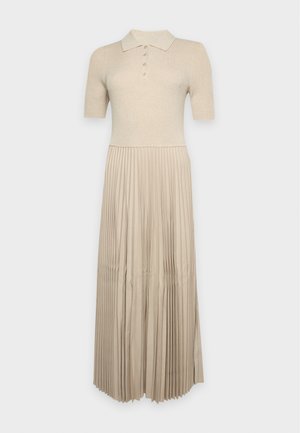 Modèle portant une robe en maille beige à manches courtes, avec une jupe midi plissée et des chaussures plates noires, devant un fond clair uni.