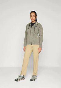 Moteris stovi su pilku užtrauktuku džinsiniu švarku, béže spalvos kelnėmis ir pilkais žygio batais prieš paprastą šviesų foną.