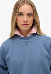 Blauwe sweatshirt met een v-hals en contrasterende stiksels, voorzien van een klein logo. Gelaagd onder een lichtroze overhemd met kraag. Glad van textuur.