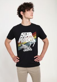 Černé bavlněné tričko s nápisem „STAR WARS“ a grafikou vesmírné lodi na pozadí vícebarevných diagonálních pruhů.