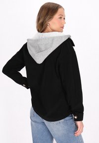 Cămașă neagră din catifea cu glugă gri, având mansete cu nasturi și o croială lejeră. Materialul este texturat, având striaturi distincte.