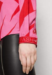 Camicia a maniche corte in rosa e rosso con un motivo audace, caratterizzata da un'etichetta rossa con la scritta "FASHION" sul polsino. Tessuto morbido, vestibilità slim.