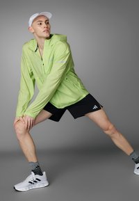 Hellgrüne, kapuzenjacke mit reflektierenden Akzenten, kombiniert mit schwarzen Shorts. Das Outfit wird mit weißen Turnschuhen und grau gestreiften Socken abgerundet.