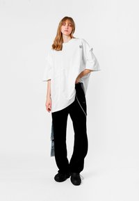 Camiseta blanca de gran tamaño con cuello redondo, costuras visibles y abertura lateral. Combinada con pantalones negros y calzado texturizado.