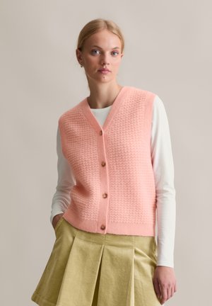 Helles rosa gestricktes Oberteil mit strukturiertem Muster, V-Ausschnitt und Holzknöpfen. Getragen über einem weißen Langarmshirt und kombiniert mit einem plissierten Rock.