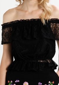 Top in pizzo nero off-the-shoulder con volants a strati e vita arricciata. Motivi floreali intricati visibili nella trama del pizzo.