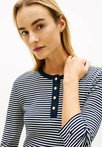 Blondynka w granatowo-białej koszulce w paski z złotymi guzikami wzdłuż przedniego plisowania, dotykająca ramienia jedną ręką.