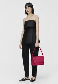Black strapless top with peplum design, paired with tailored black pants. Model holds a bright pink handbag with embossed "TOUS" logo.