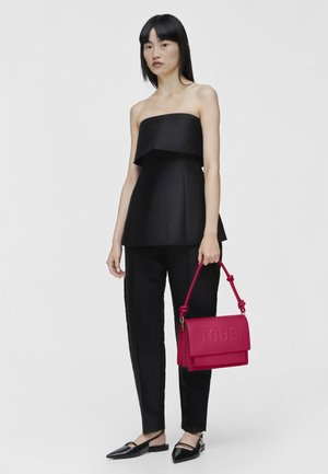Haut noir sans bretelles avec un design peplum, associé à un pantalon noir ajusté. Le mannequin tient un sac à main rose vif avec le logo en relief "TOUS".