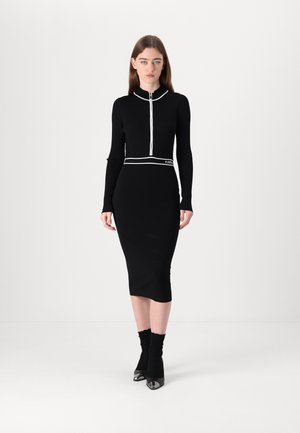 Black ribbed dress with a high neckline, featuring a front zipper. White accents at the collar and waist, paired with black fitted socks and heeled shoes.