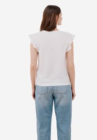 Top bianco a maniche corte con dettagli a volants, abbinato a jeans chiari a vita alta. Tessuto morbido, vestibilità casual, design minimale.