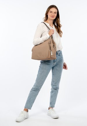Beige strukturierte Tote-Tasche mit einem Reißverschluss, dekorativem Quasten und einem geprägten Logo. Lässig kombiniert mit hellblauen Jeans und weißen Sneakers.