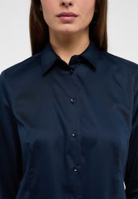 Granatowa koszula zapinana na guziki, wykonana z gładkiego materiału, z kołnierzykiem w kształcie szpica i ośmioma pasującymi guzikami z przodu. Prosty i elegancki design.