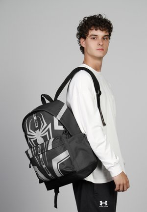 Sac à dos noir et gris présentant un motif de toile d'araignée, en tissu texturé, avec une poche avant et des bretelles ajustables, porté par une personne.