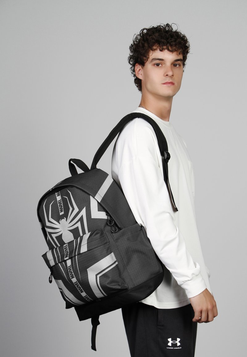 Sac à dos noir et gris présentant un motif de toile d'araignée, en tissu texturé, avec une poche avant et des bretelles ajustables, porté par une personne.