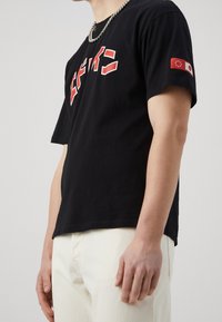 Černé bavlněné tričko s výrazným červeným nápisem a vlajkami Singapuru a Japonska na rukávu. Krátké rukávy a standardní střih.