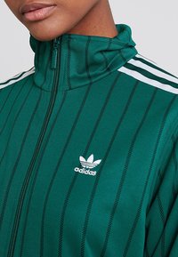 Grön zip-up jacka med vertikala svarta ränder. Har en hög krage och vitt Adidas-logotyp, accentuerad med vita ärmränder.