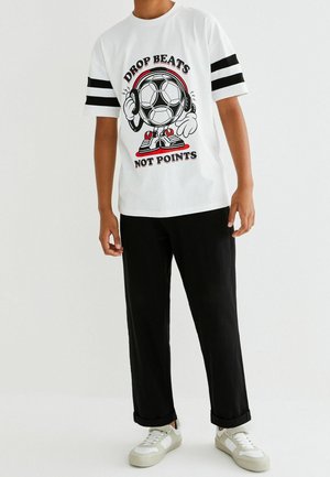 Personne portant un t-shirt blanc avec des rayures noires sur les manches montrant un ballon de football dessin animé avec des écouteurs et le texte "Drop beats not points."