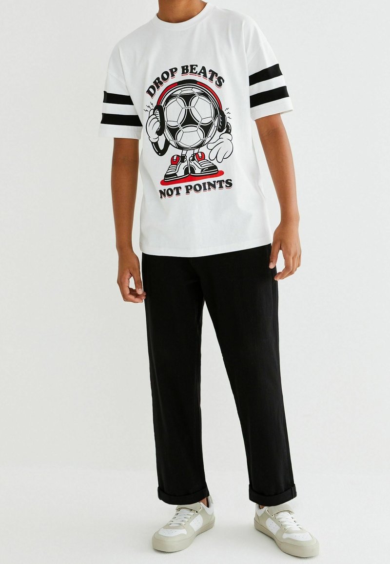 Personne portant un t-shirt blanc avec des rayures noires sur les manches montrant un ballon de football dessin animé avec des écouteurs et le texte "Drop beats not points."